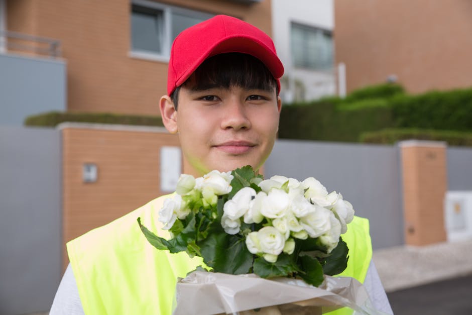 A young male florist assistant wearing a red cap and a neon yellow safety vest holds a bouquet of fresh white roses with green foliage, wrapped in white paper. He stands outdoors in front of modern re