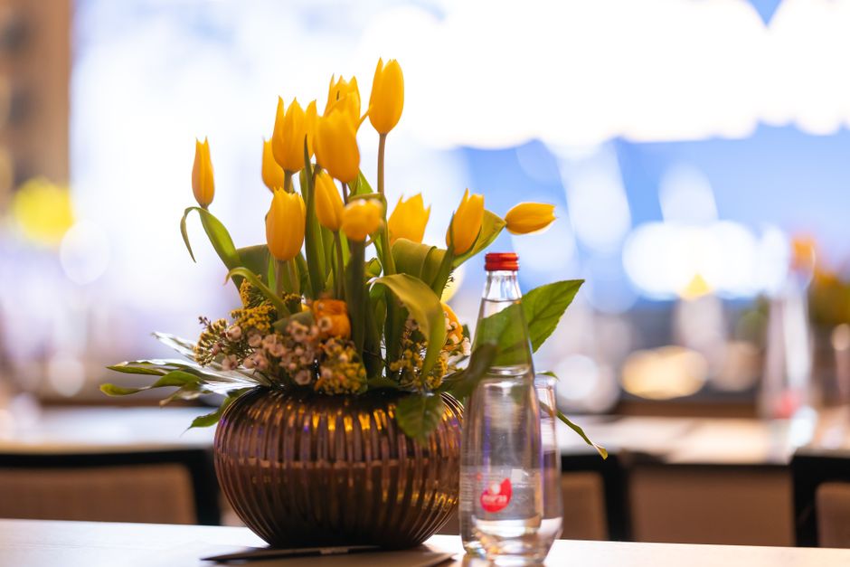 A floral arrangement featuring bright yellow tulips with closed and partially open petals, accompanied by smaller sprigs of pink baby's breath and green foliage, is arranged in a rounded, ribbed amber