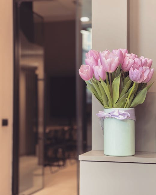 A bouquet of pink tulips with fresh, vibrant petals and green stems arranged in a light green ceramic vase wrapped with a pale purple ribbon. The vase is placed on a white surface inside an indoor set