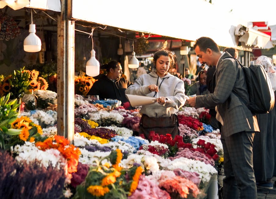 A vibrant outdoor flower market scene with fresh flowers arranged in colorful bouquets and bunches, including sunflowers, roses, chrysanthemums, and daisies in shades of yellow, white, pink, red, purp