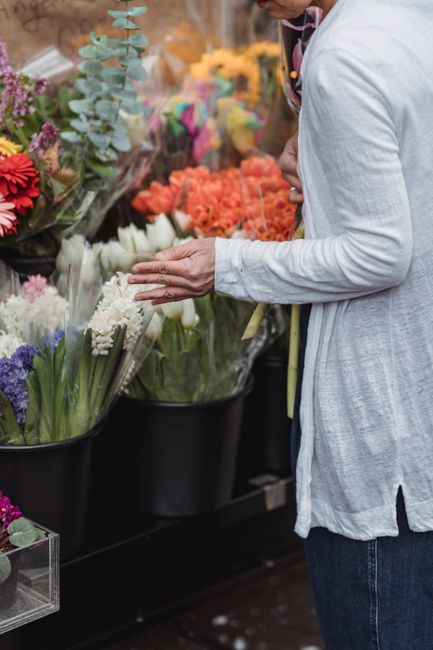 A person in a white long-sleeved shirt selecting fresh flowers from a display at a flower shop, with tulips, hyacinths, and other blooms arranged in black containers. The flowers include white hyacint
