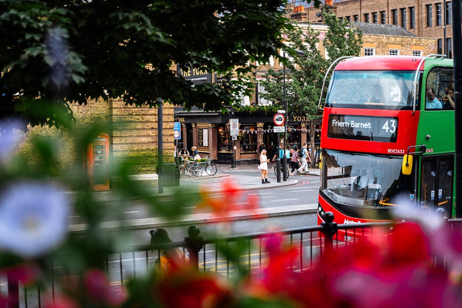 A view of a busy street in New Barnet, showing a double-decker bus with a red and green livery and the route number 43, labeled 'Friern Barnet.' In the foreground, blurred colorful flowers and greener