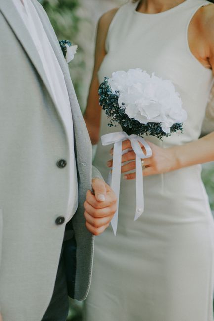 A close-up of a bride holding a small bouquet of white hydrangeas, accented with dark green foliage, tied with a white satin ribbon. The bride is wearing a sleeveless ivory wedding dress, and her hand
