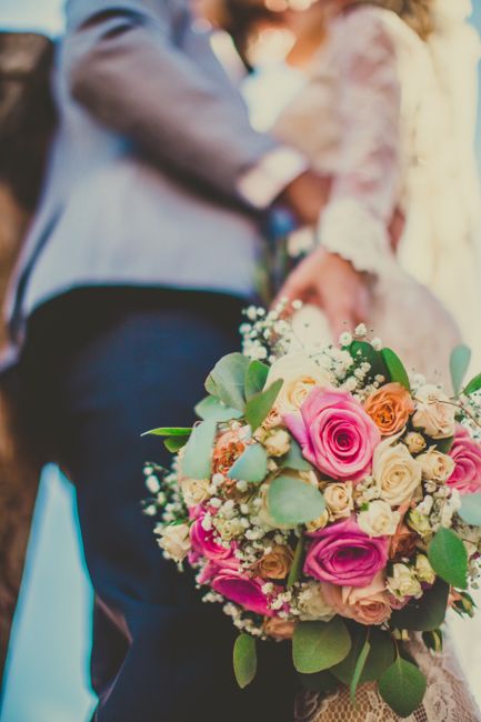 A close-up view of a floral bouquet featuring pink roses, cream roses, and small white filler flowers, accented with eucalyptus leaves. The bouquet is lush and round, wrapped in soft lace and tied wit