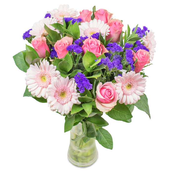 This striking floral arrangement, artfully composed by a New Barnet florist, stands in a clear glass vase set against a bright, unadorned backdrop. At its heart are seven to nine pink roses, their petals arranged in classic, tightly furled spirals with some just beginning to bloom, hinting at secrets and new beginnings often celebrated in New Barnet homes. Encircling them are five to seven pale pink gerbera daisies, their broad, daisy faces and soft, nearly white centres drawing light like the open spaces of Oakhill Woods. Threaded throughout, vivid clusters of purple statice or limonium add depth, texture, and a vibrant contrast-much like bursts of colour one might find at the weekly New Barnet Farmers' Market. A generous base of deep green foliage provides natural volume and a sense of lushness, enhancing the bouquet's visual freshness and the idea of abundance found in neighbourhood gardens. The slender transparent vase allows the eye to follow each stem down into clear water, adding to the arrangement's sense of clean, deliberate beauty. Perfect for birthdays, anniversaries, Mother's Day, or as a thoughtful decorative accent, this bouquet expresses joy and affection, infusing local homes and offices with the timeless elegance of expertly arranged blooms.